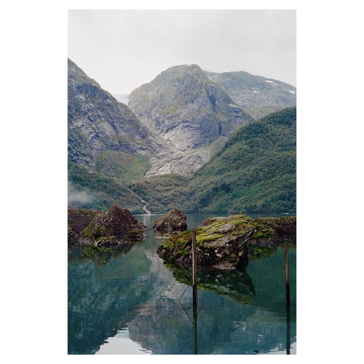A body of water with mossy rocks and wooden posts reflects mountains and a cloudy sky.