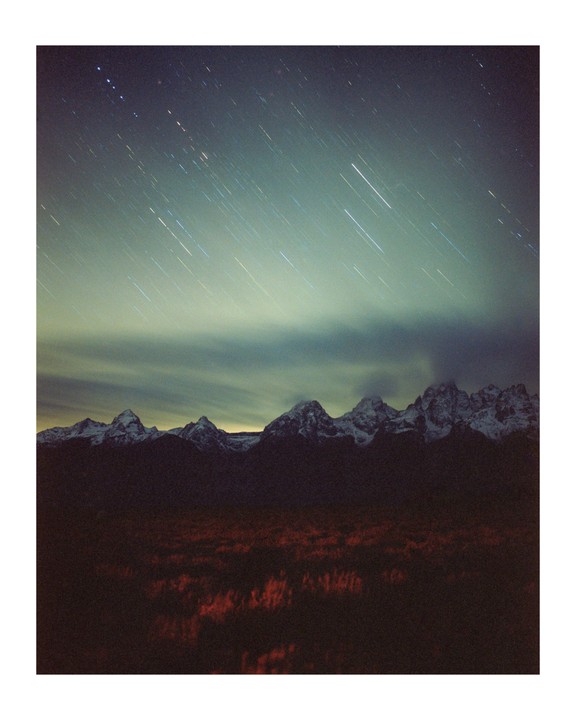 Star trails are visible above a mountain range and a field of red foliage at night.