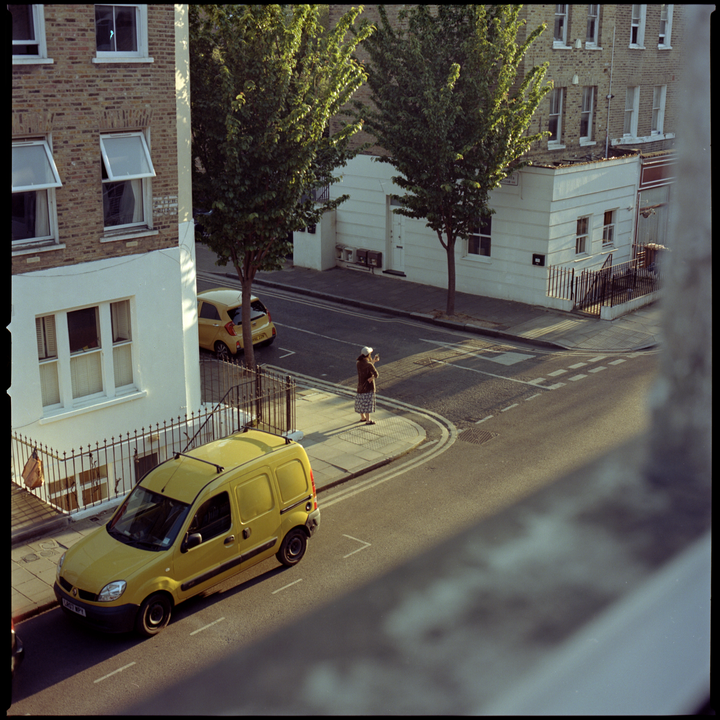 A woman stands on a street corner next to a yellow van, with buildings and trees in the background.