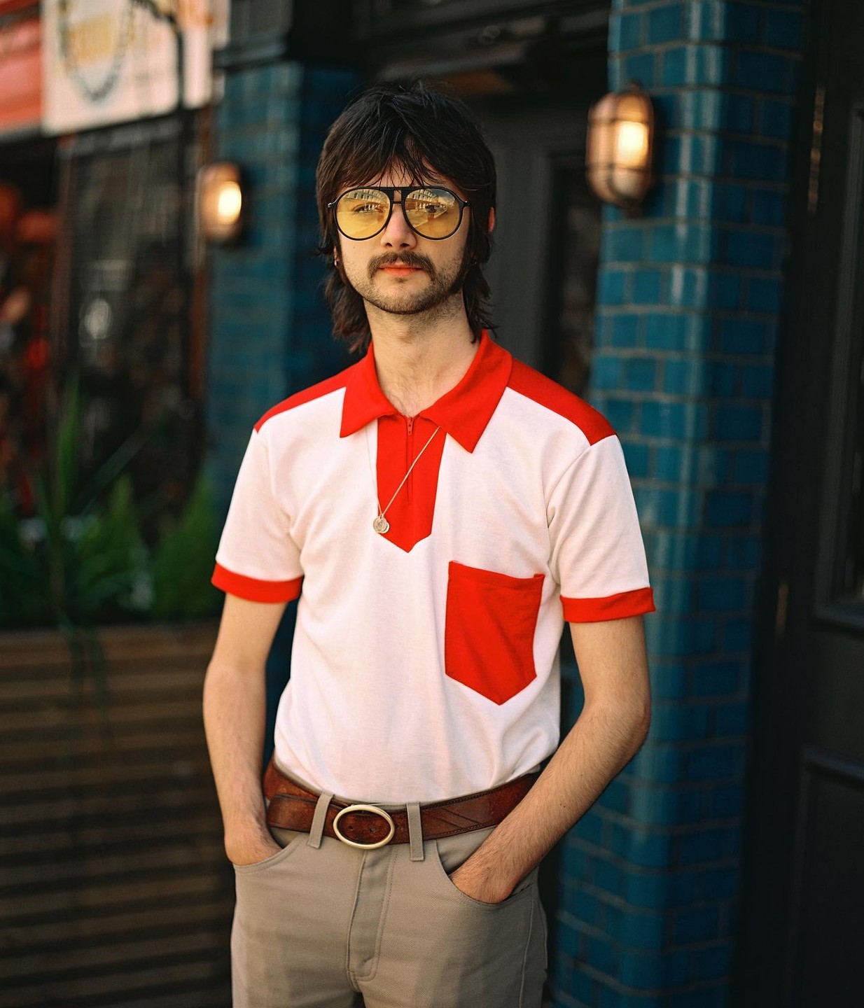 A man with a mustache and yellow-tinted glasses stands with his hands in his pockets.