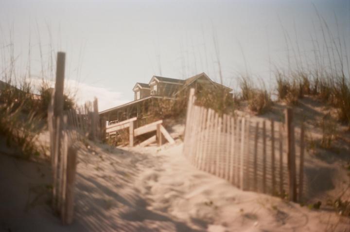 A wooden fence and a path lead through sand dunes towards a house.