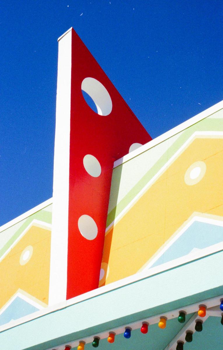 A red triangular architectural detail with white circles rises against a blue sky, next to a yellow and blue building facade with colorful lights.