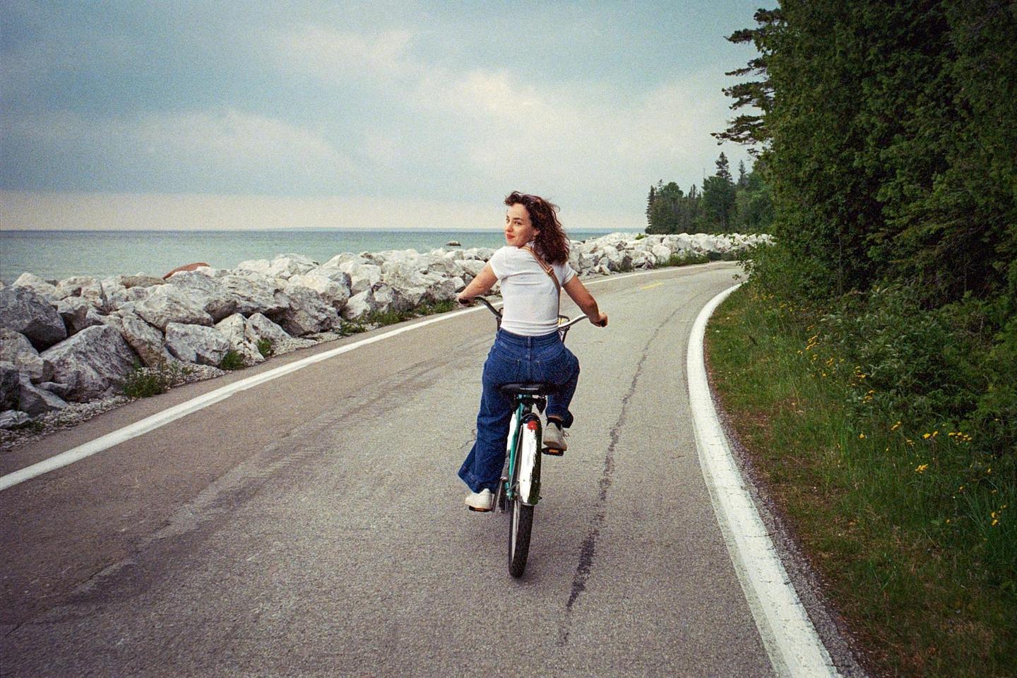 A woman on a bicycle looks back at the camera while riding on a paved road next to a rocky shore and ocean.