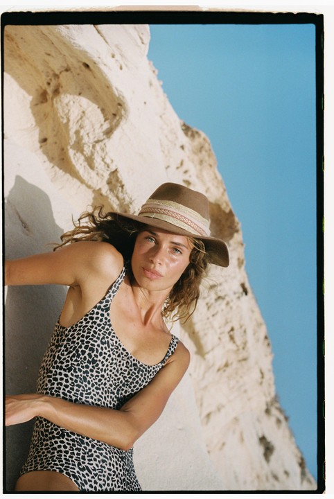 A woman in a leopard print swimsuit and a hat poses against a light-colored rock formation under a clear blue sky.