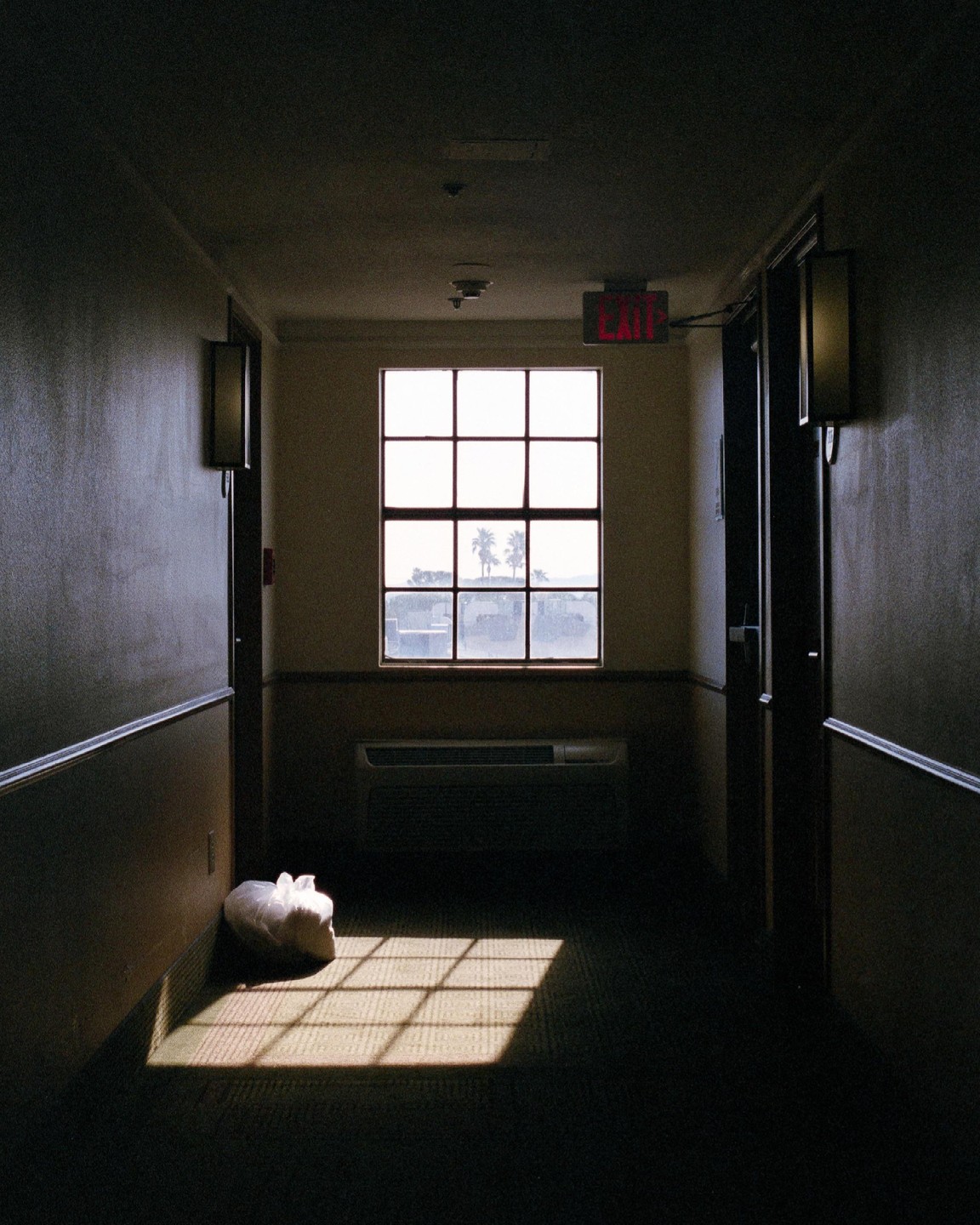 A long, dark hotel hallway leads to a bright window with an exit sign above it.