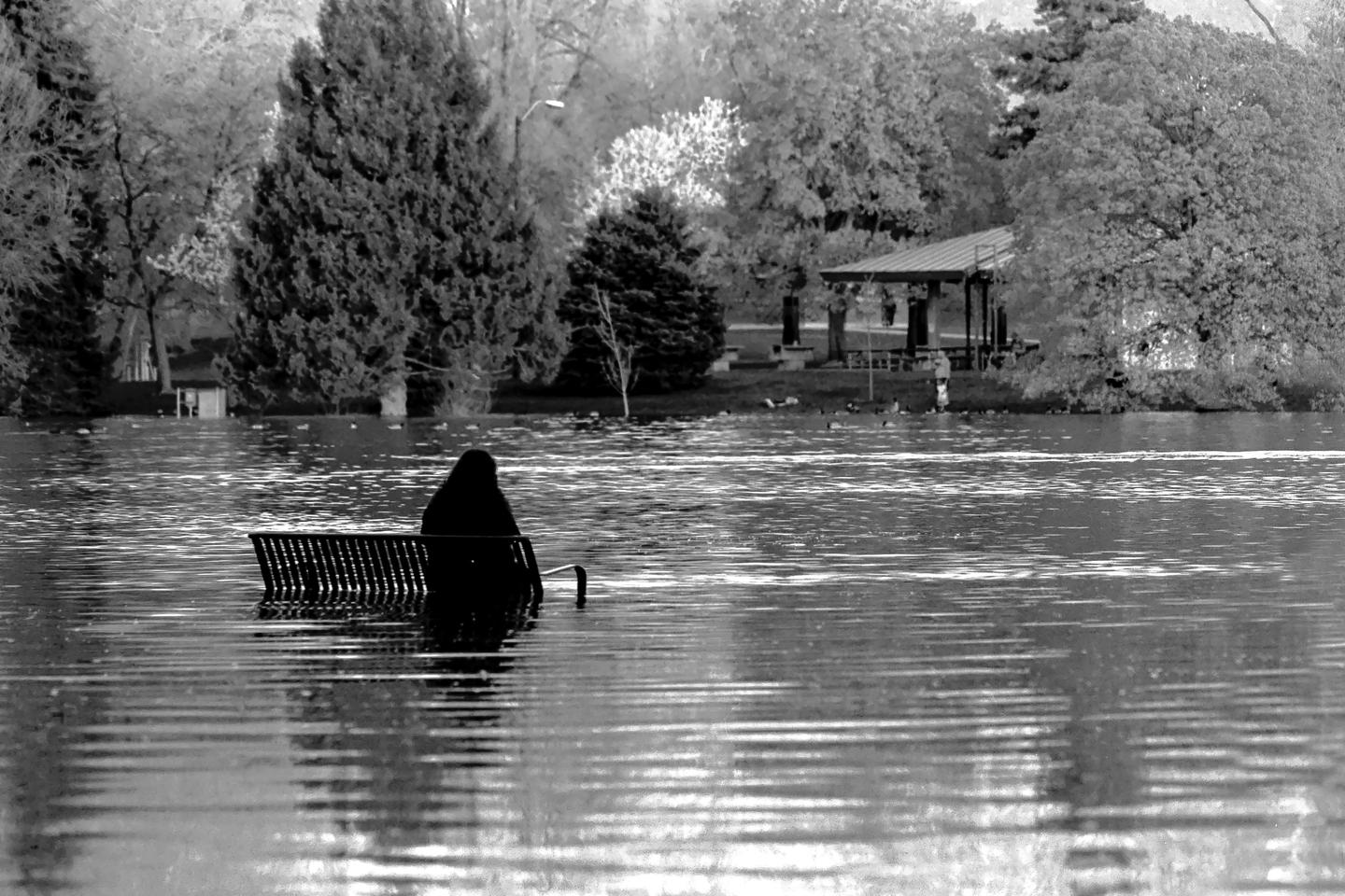 A person sits on a partially submerged park bench in a flooded park with trees and a gazebo in the background.
