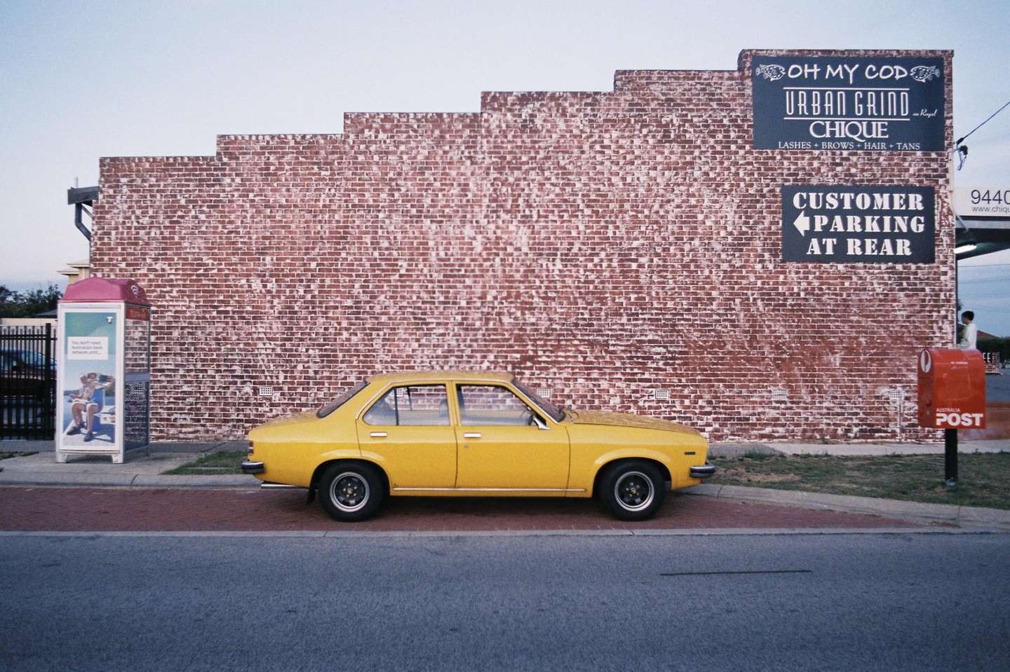 A yellow car is parked on a street next to a brick wall with signs and a red mailbox.