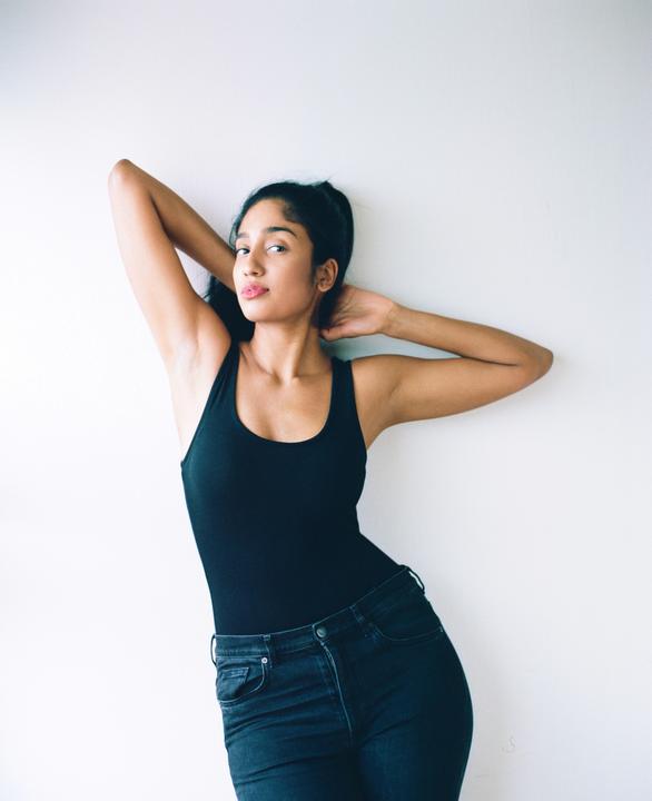 A woman in a black tank top and jeans poses against a white wall with her arms behind her head.