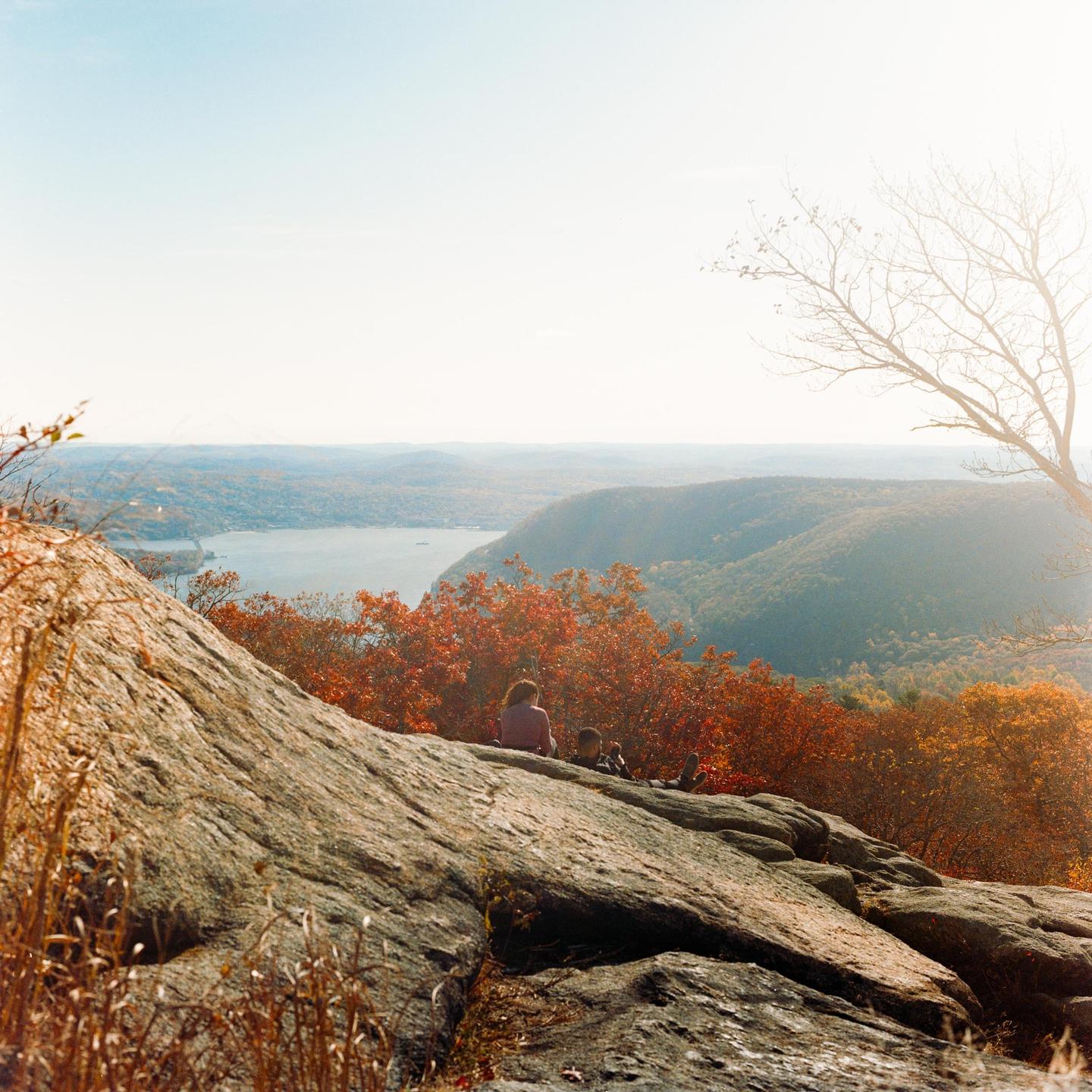 A woman and child sit on a rocky outcrop overlooking a river and mountains covered in autumn foliage.