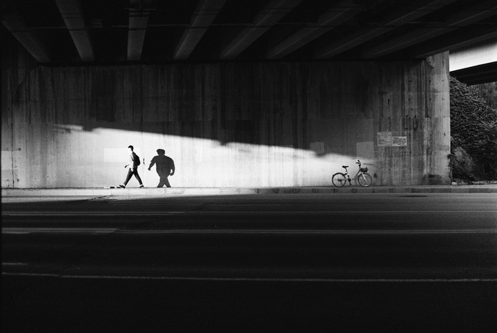 A man walks along a sunlit wall under a bridge, casting a large shadow, with a bicycle parked nearby.