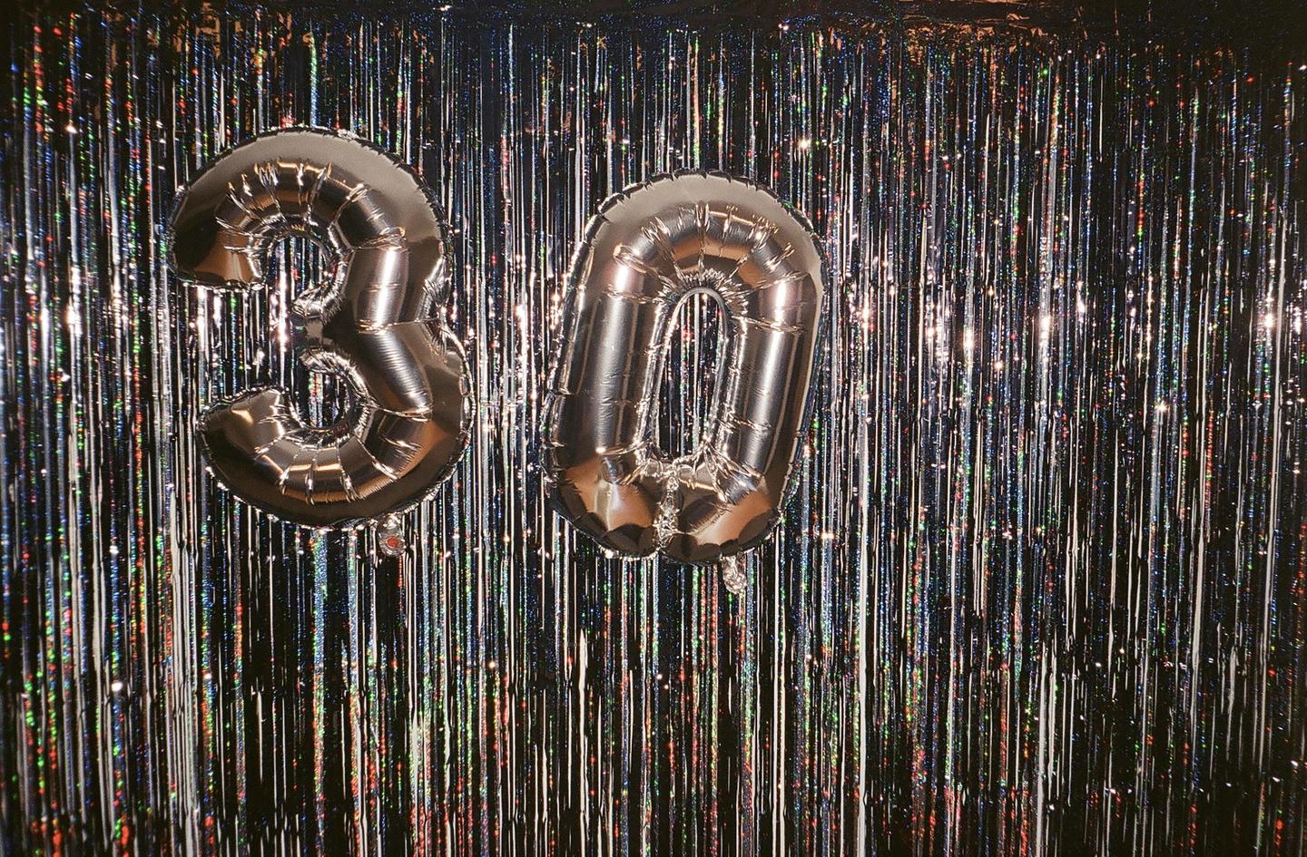 Two silver balloons shaped like the numbers three and zero float in front of a shimmering curtain.