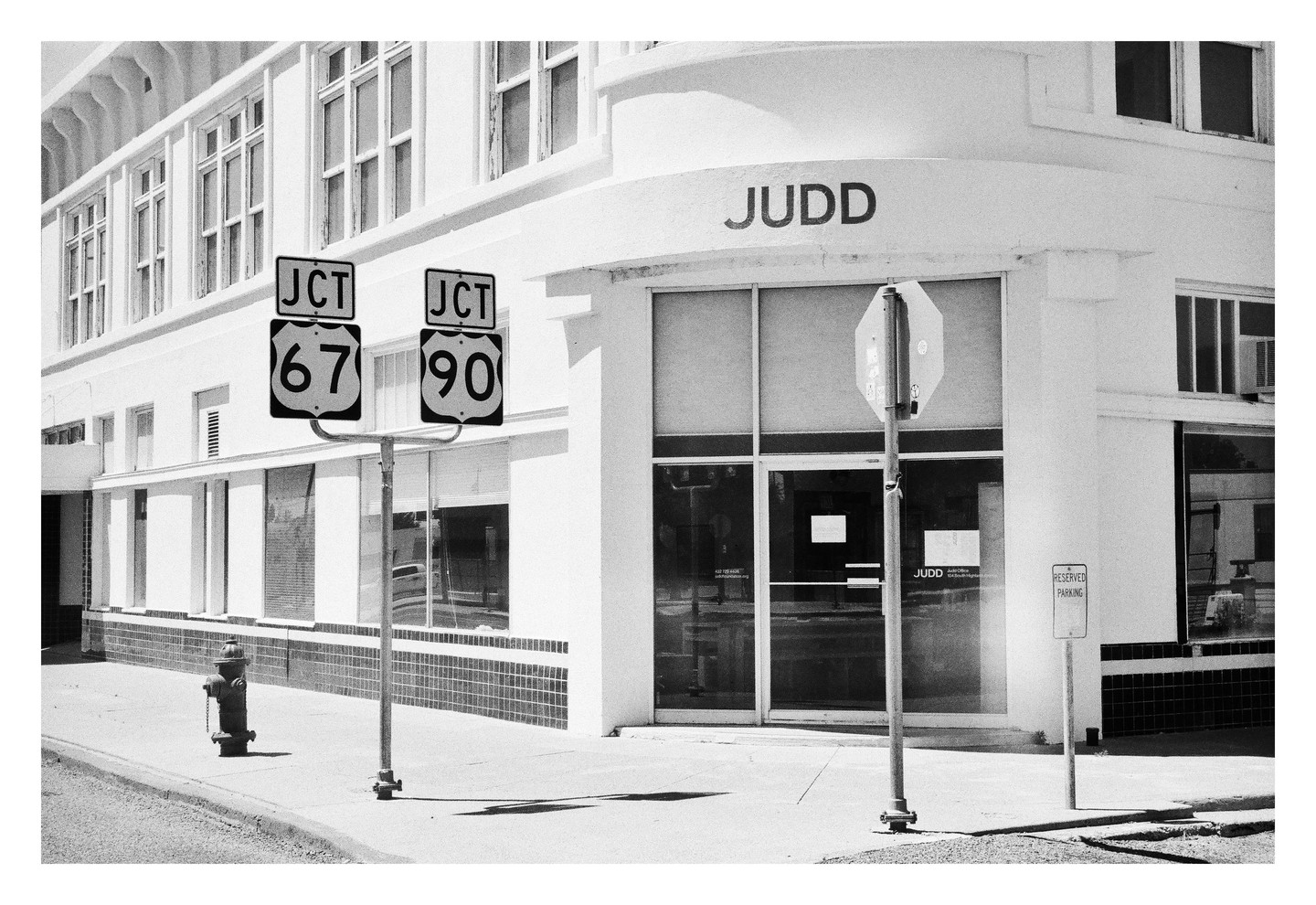 A street corner features a white building with "JUDD" written on it, two highway signs for routes 67 and 90, a fire hydrant, and a "reserved parking" sign.