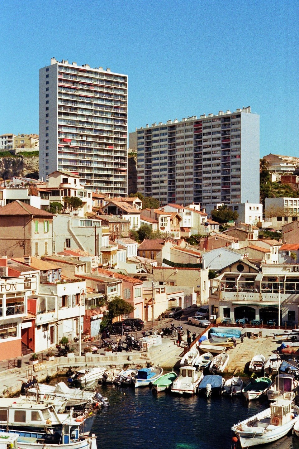 A small harbor with many boats is surrounded by buildings and two large apartment blocks under a clear sky.