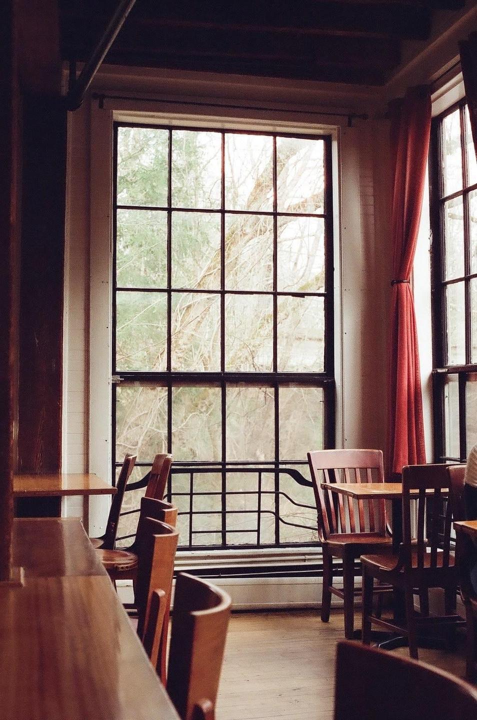 A large window with a grid pattern looks out onto trees, with wooden tables and chairs in the foreground.