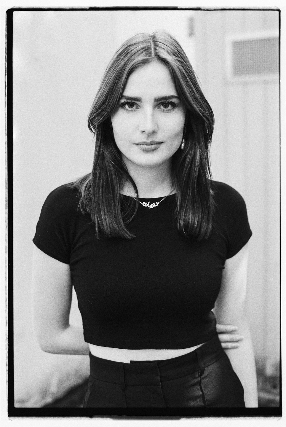 A woman with long hair and a necklace stands with her arms behind her back.