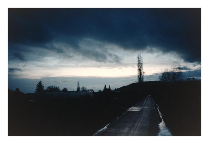 A dark road with puddles leads into the distance under a dramatic, cloudy sky at dusk.