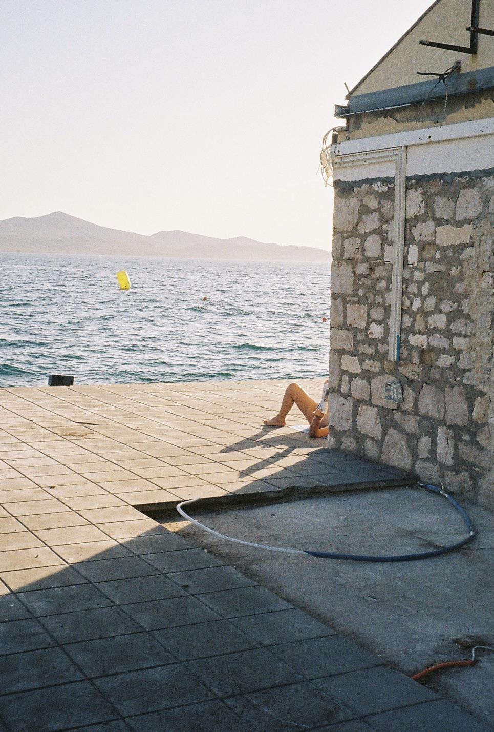 A person is relaxing on a tiled pier next to a stone building, with the sea and mountains in the background.