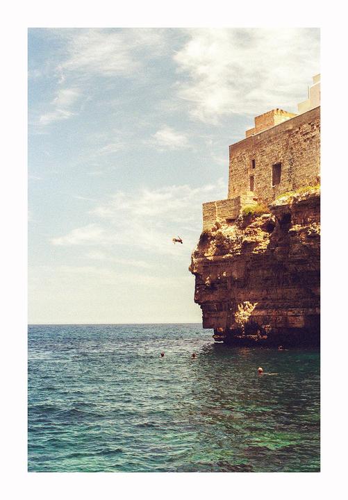 A person dives from a cliff next to a stone building into the ocean.