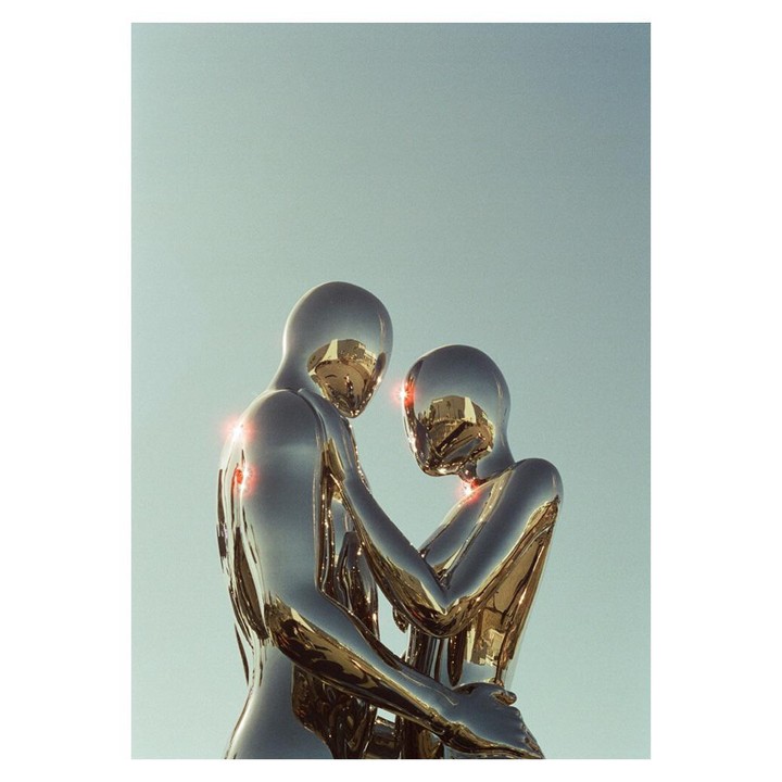 Two metallic, humanoid figures embrace under a clear sky.