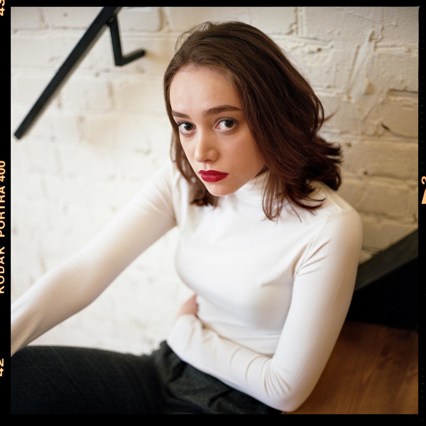A woman with red lipstick and a white turtleneck sits on a wooden floor next to a white brick wall with a black railing.