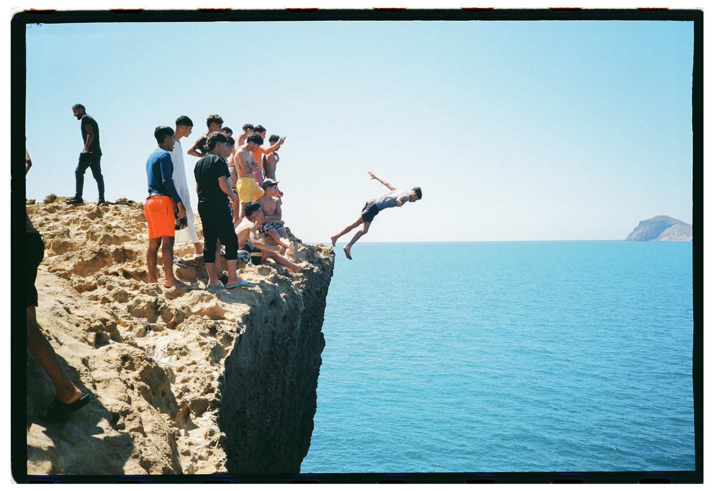 A group of men stand on a rocky cliff as one man dives into the blue ocean.
