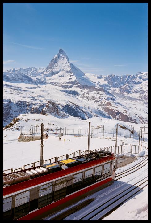 A red train is on tracks in a snowy mountain landscape with a prominent peak in the background.