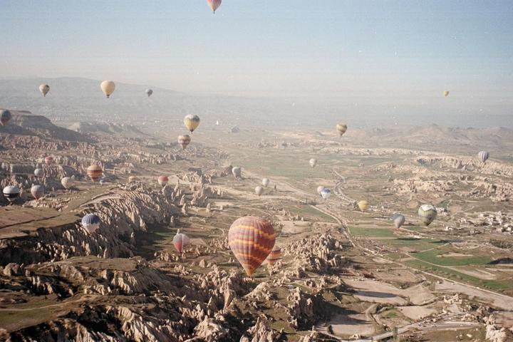 Hot air balloons float above a rugged, arid landscape with some green fields and a small town.