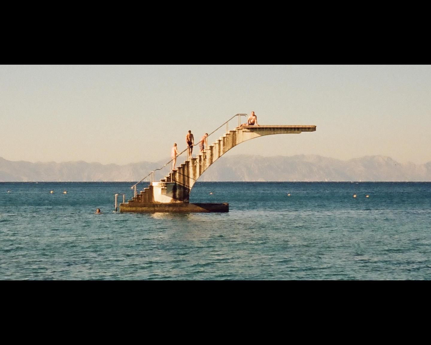 Several people are on and around a diving board structure in the middle of a body of water with mountains in the distance.