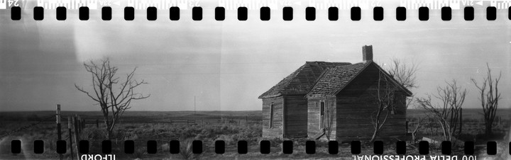 An abandoned wooden house with a chimney stands in a desolate field with bare trees under a cloudy sky.