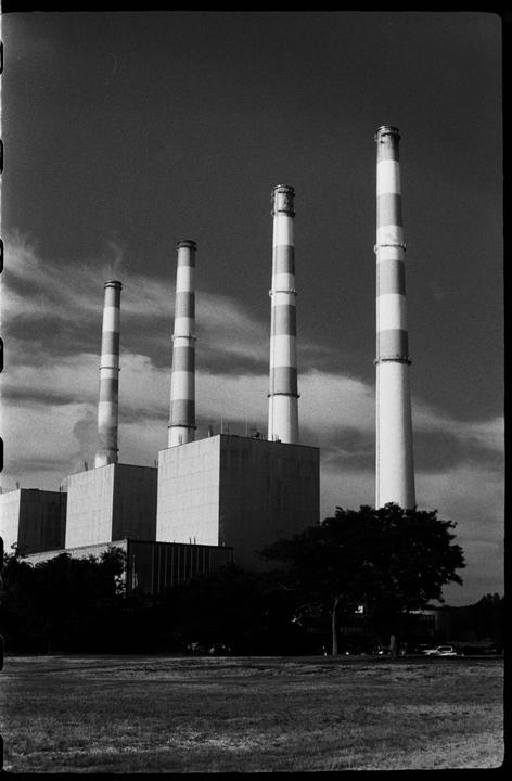 Four striped smokestacks rise above a power plant with a grassy field and trees in the foreground.