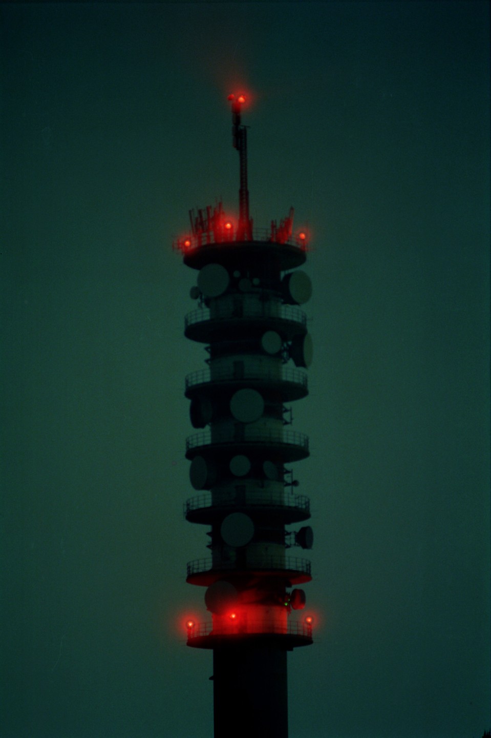 A tall communication tower with multiple red lights glows against a dark, greenish-blue sky.