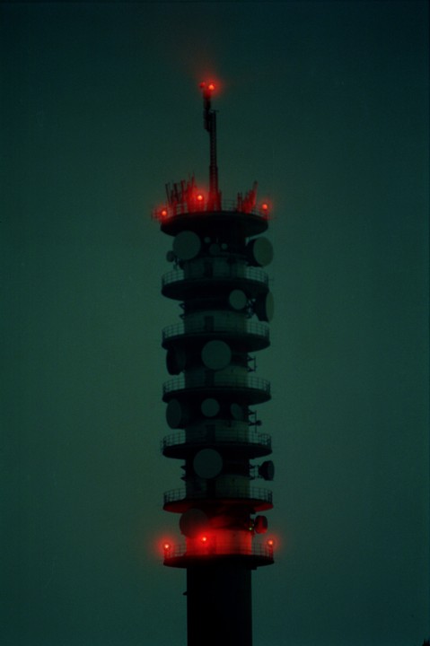 A tall communication tower with multiple red lights glows against a dark, greenish-blue sky.