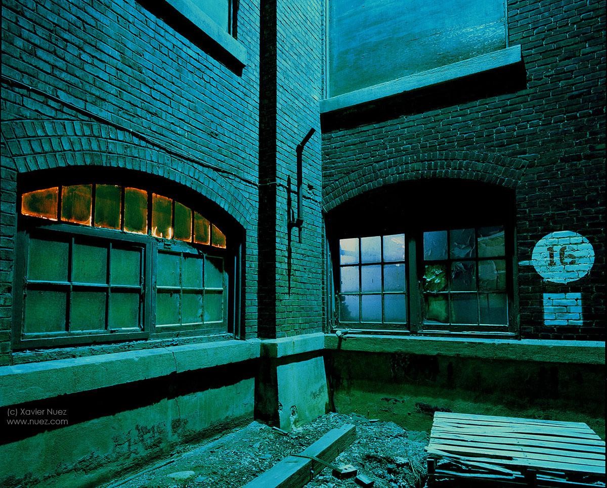 Two arched windows are set into a brick building, one with an orange glow and the other reflecting blue light, with a number 16 painted on the wall.