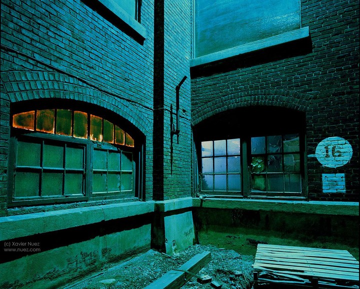 Two arched windows are set into a brick building, one with an orange glow and the other reflecting blue light, with a number 16 painted on the wall.