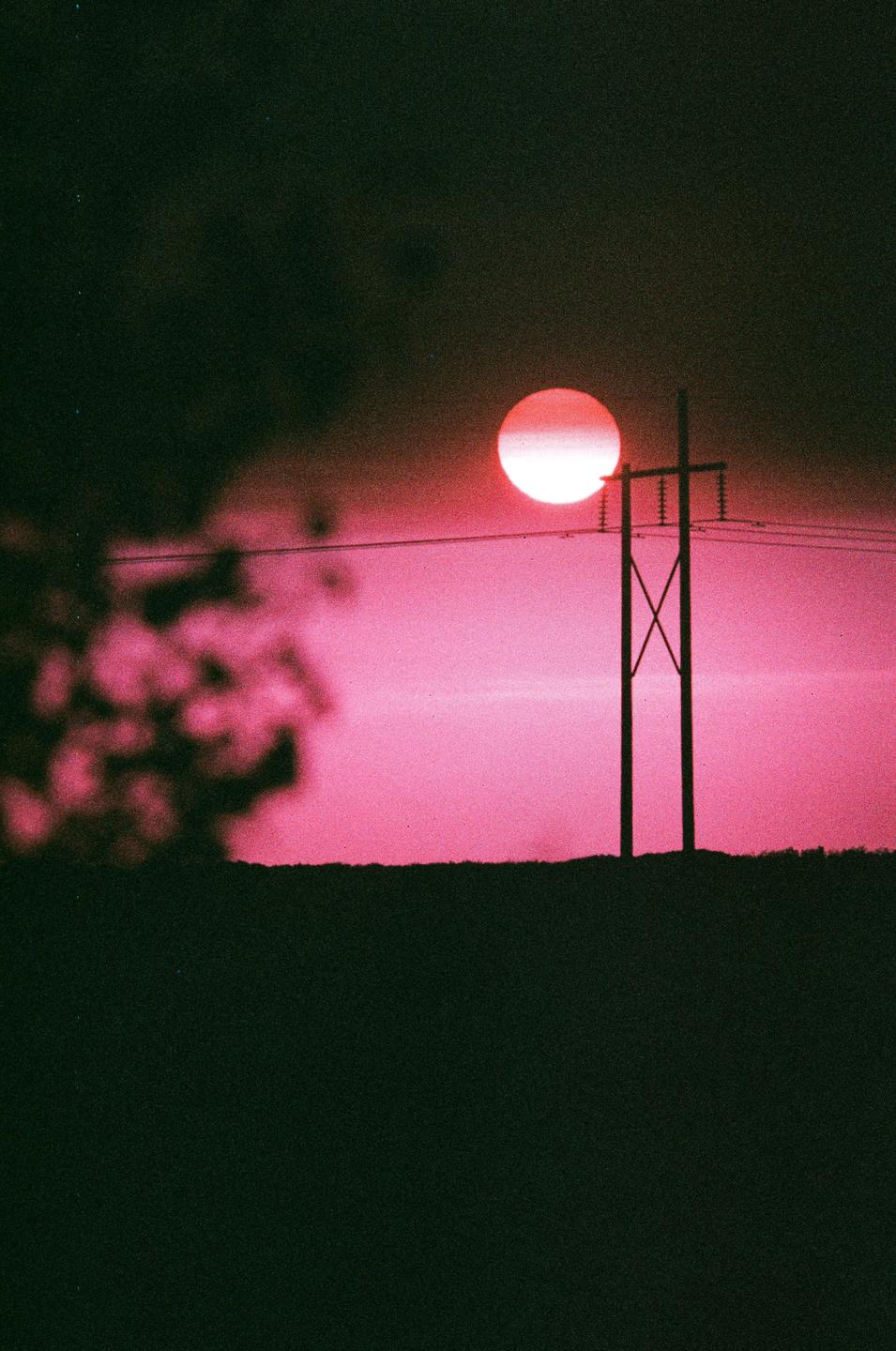 A large red sun is setting behind a power line and silhouetted landscape.