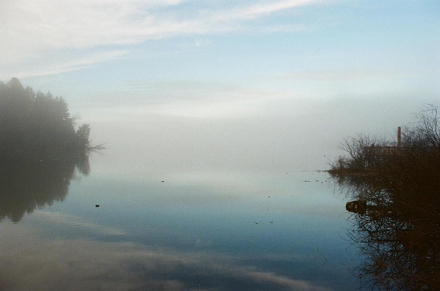 A body of water reflects the sky and trees on a hazy day, with a building and chimney visible on the right bank.