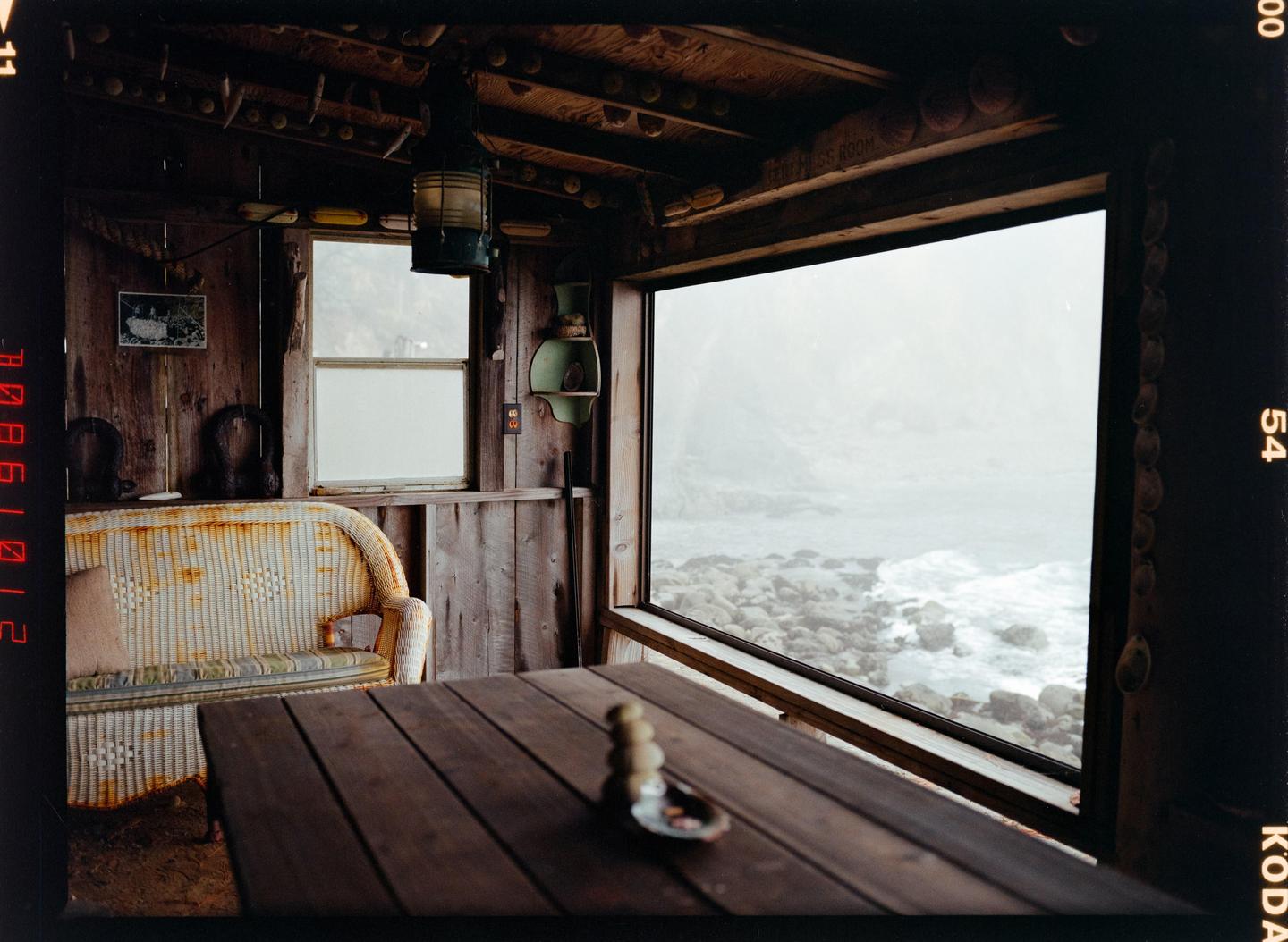 A rustic wooden room with a large window overlooks a rocky, misty coastline.