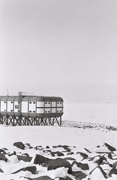 A research station stands on stilts in a snowy, rocky landscape under a bright sky.