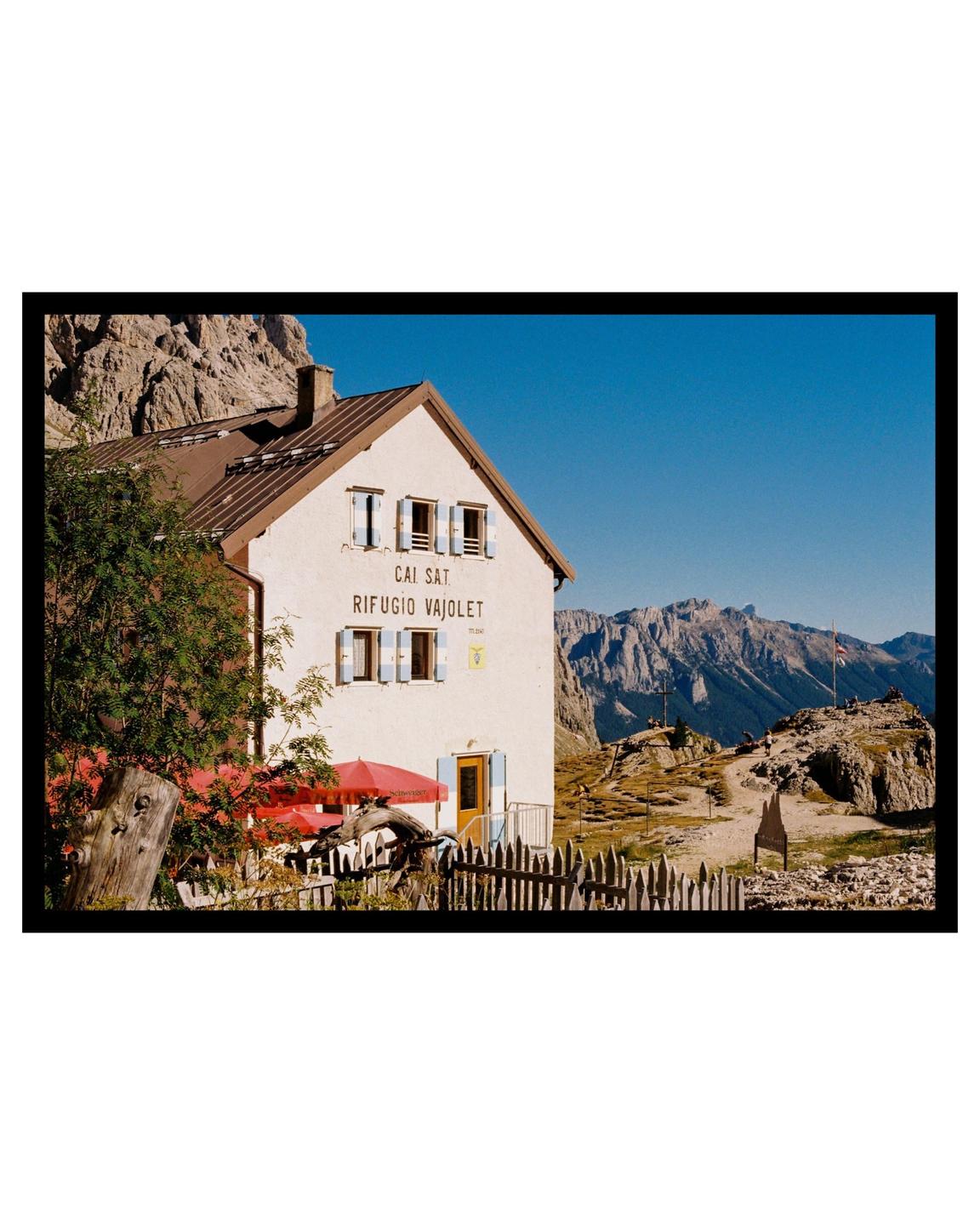 A white mountain hut with a red umbrella and a wooden fence stands against a backdrop of rocky mountains and a clear blue sky.