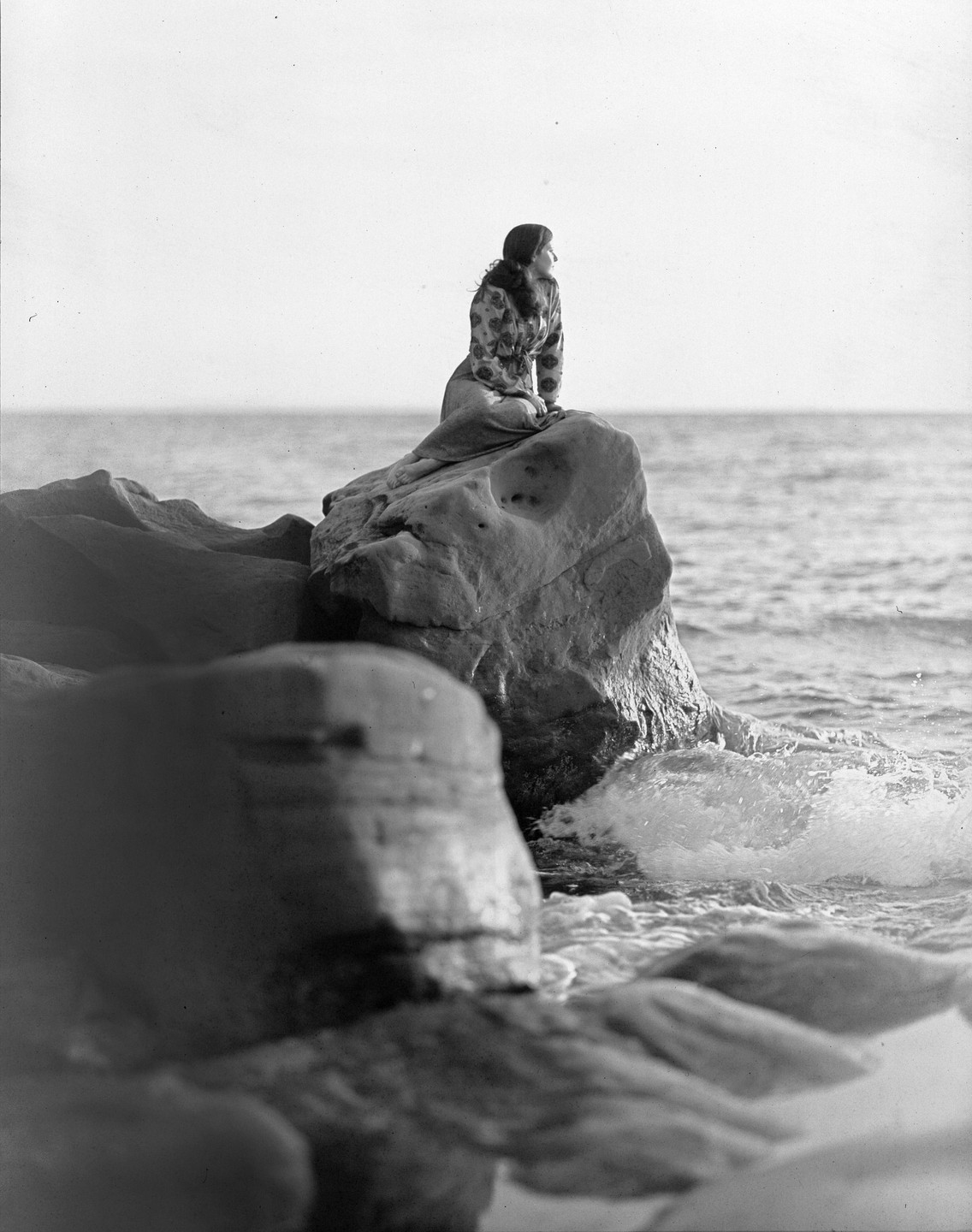 A woman sits on a large rock by the ocean, looking out at the water.