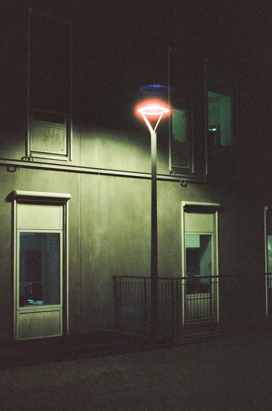 A street light with a red glow and a blue halo stands in front of a building with windows at night.