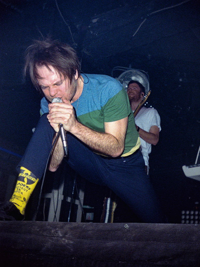 A man with messy hair sings into a microphone on stage, wearing a blue and green shirt, dark pants, and yellow socks with black lettering.