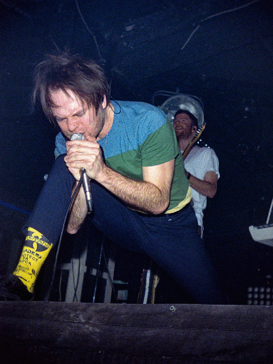 A man with messy hair sings into a microphone on stage, wearing a blue and green shirt, dark pants, and yellow socks with black lettering.