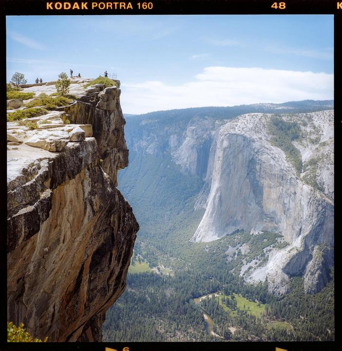 A group of people stand on a cliff overlooking a valley with a river and mountains.