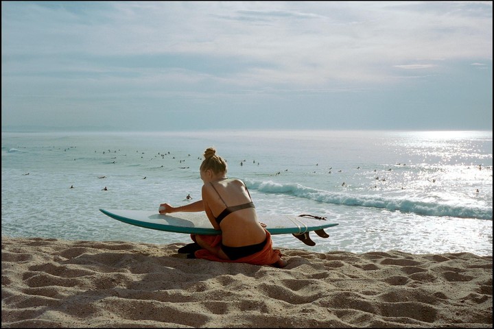 A woman sits on a sandy beach, waxing a surfboard while looking out at the ocean filled with surfers.