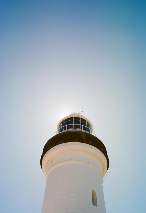 A white lighthouse with a dark top is seen from below against a bright blue sky.