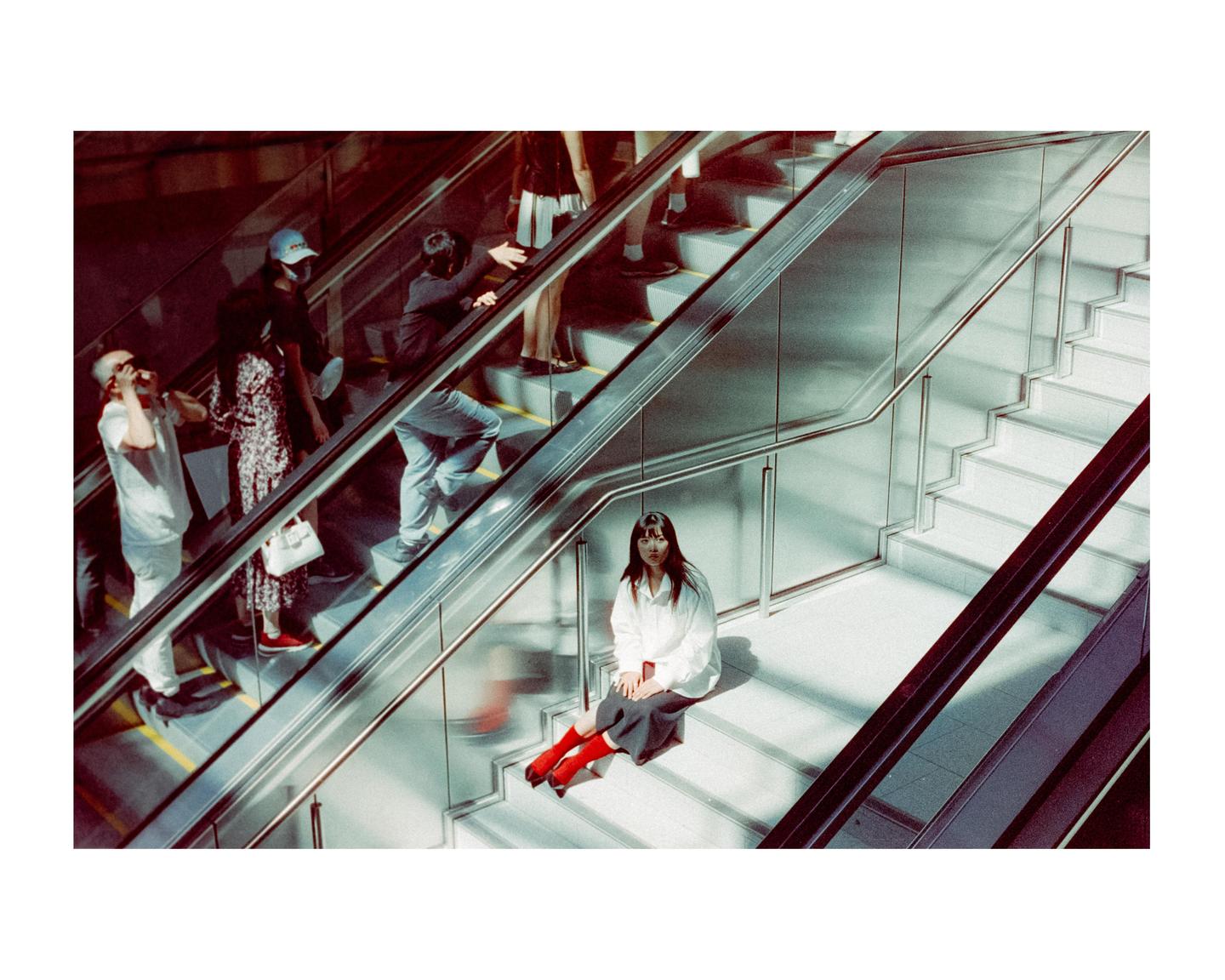 A woman sits on a staircase next to an escalator with people riding it.