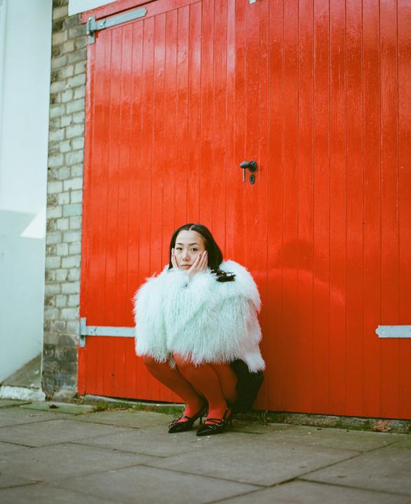 A woman in a fluffy coat and red tights squats in front of a red garage door.