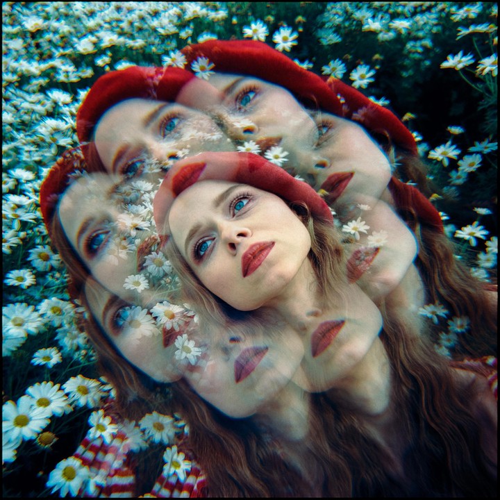 A woman with long hair and a red beret is shown in a multiple exposure, lying in a field of white daisies.