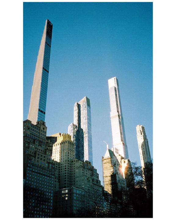 A low-angle view shows several skyscrapers towering over older buildings and trees against a clear blue sky.